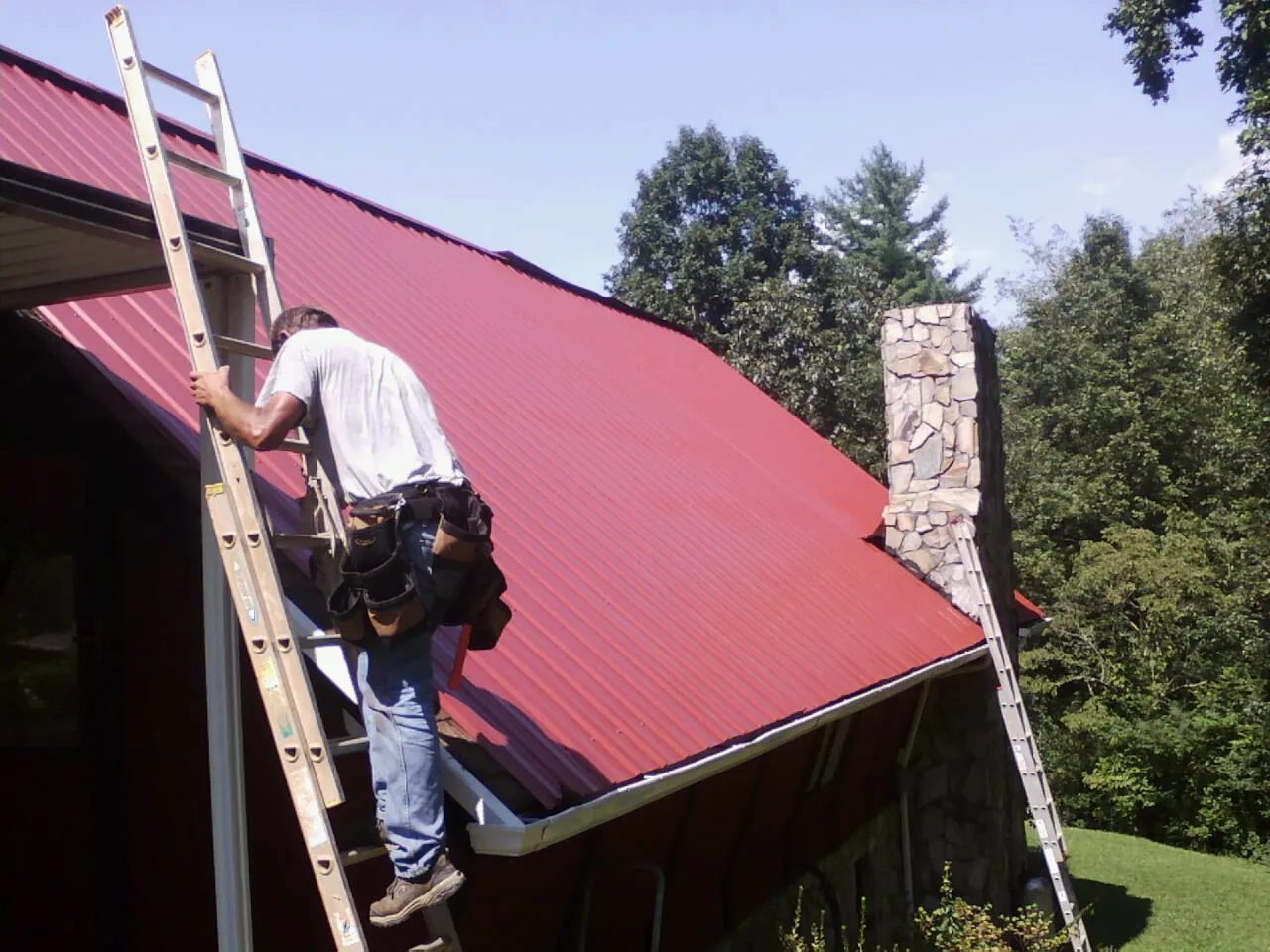 Roofer on a ladder beside a red metal roof and stone chimney — Soffit & Fascia Repair by Roofing Brothers in Prairie Lea